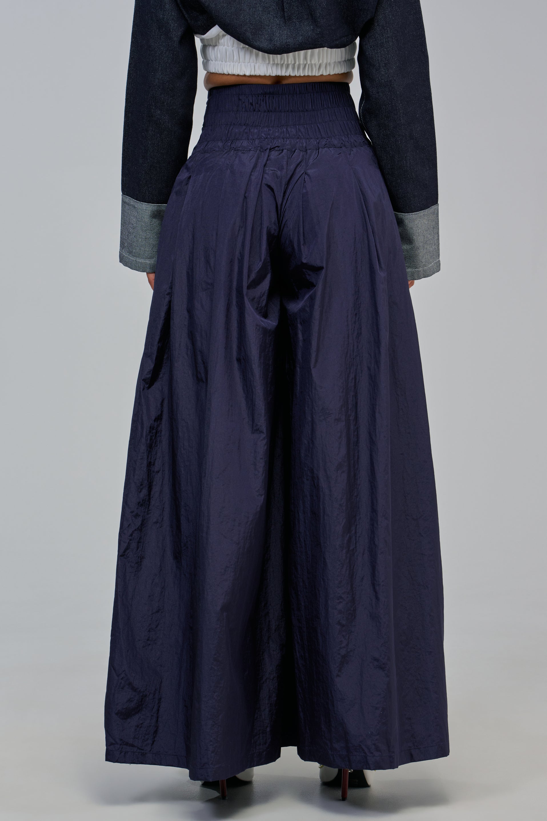 A person wearing the DANCE THE NIGHT AWAY PALAZZO PANT IN NAVY, featuring a high-waisted, wide-leg silhouette with a gathered waistband and shiny fabric, paired with a cropped dark jacket, creates a stylish sporty streetwear look. The photo is taken from behind to highlight the modern loose fit of the palazzo pants.