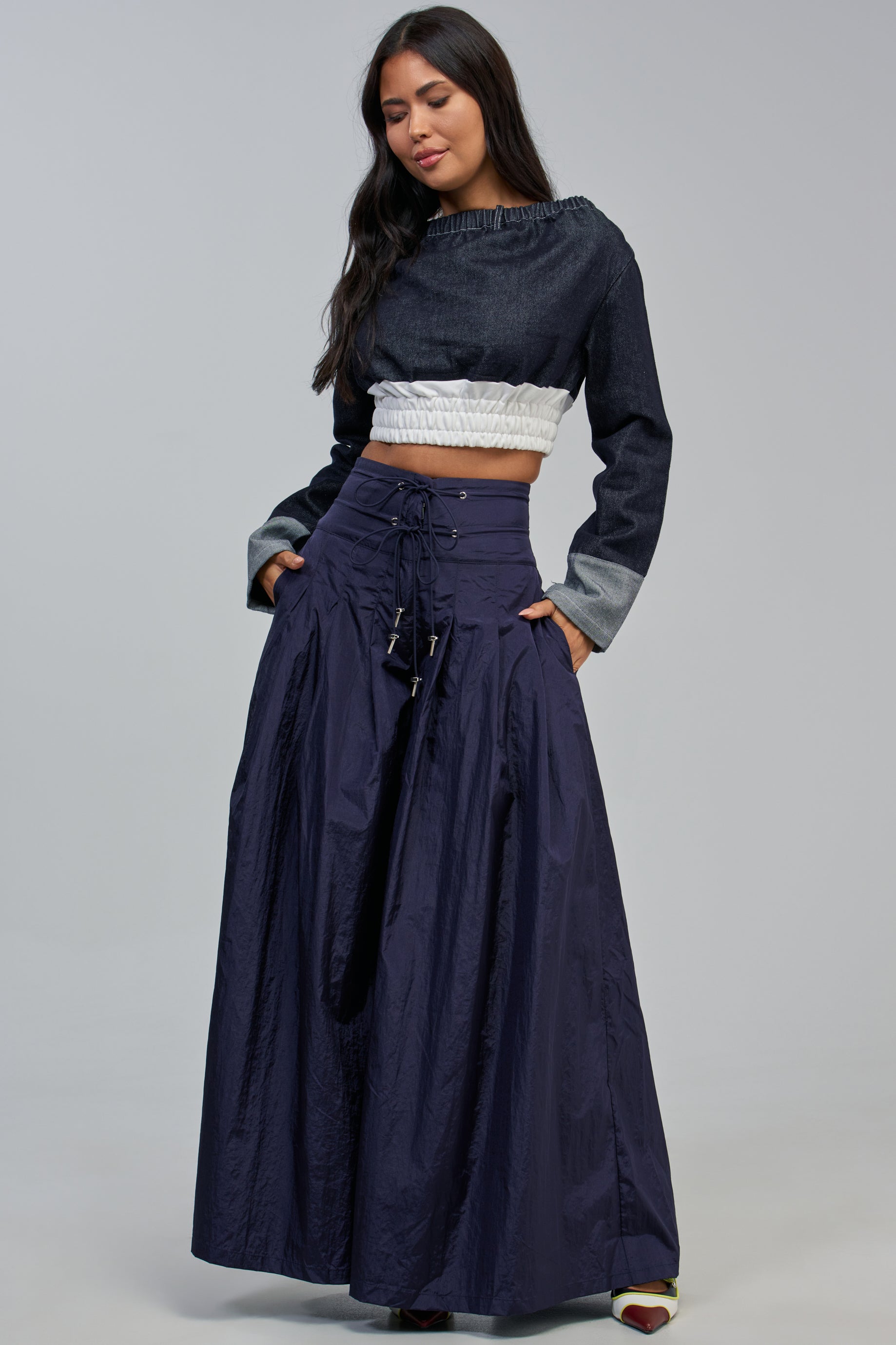 A woman stands and looks down, wearing the DANCE THE NIGHT AWAY PALAZZO PANT IN NAVY styled with a dark long-sleeved crop top featuring a white ruffled midsection, and open-toed shoes, set against a plain gray background.