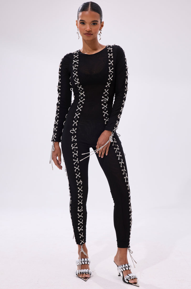 A woman exudes confidence in the LACE IT UP MESH JUMPSUIT WITH RHINESTONE DETAIL, featuring white crisscross lace-up accents and subtle rhinestones—an ideal choice for a chic nightlife look. She completes the outfit with white strappy heels and a sleek pulled-back hairstyle.