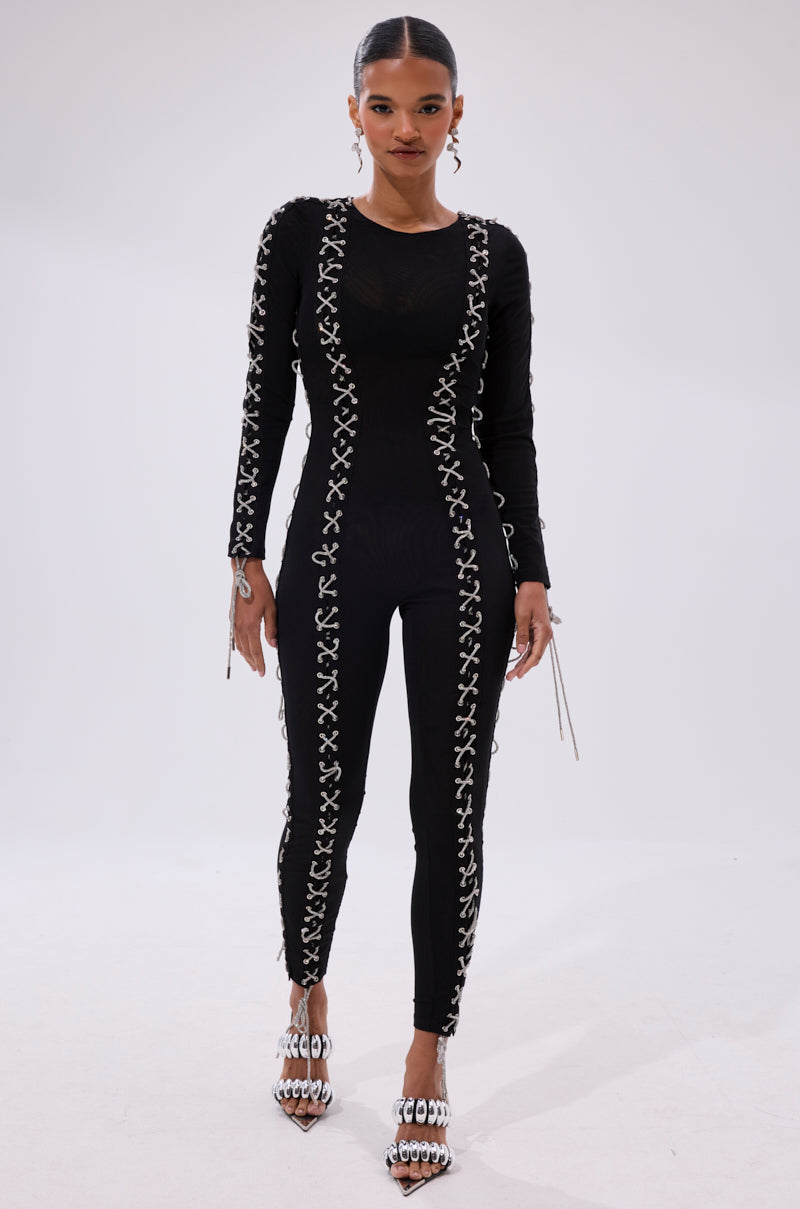 A woman faces forward in the LACE IT UP MESH JUMPSUIT WITH RHINESTONE DETAIL, a fitted black mesh piece with white lace-up accents, paired with white heeled sandals adorned with silver chain details.