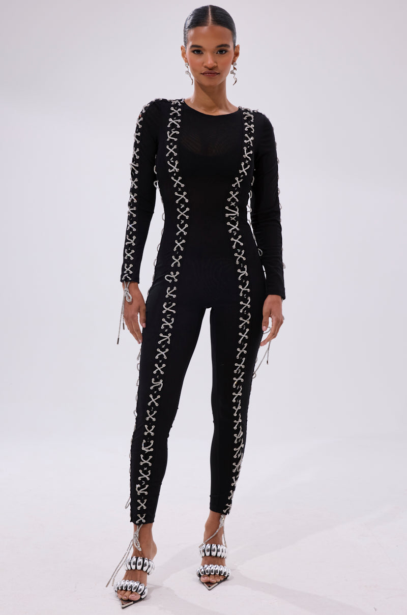 A woman faces forward in the LACE IT UP MESH JUMPSUIT WITH RHINESTONE DETAIL, a fitted black mesh style with white X-shaped stitching and lace-up accents, styled with silver heels and hoop earrings against a plain light backdrop.