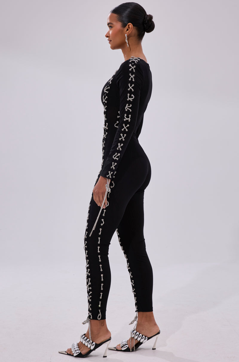 A woman stands in profile wearing the LACE IT UP MESH JUMPSUIT WITH RHINESTONE DETAIL, styled with a sleek bun, large hoop earrings, and heeled sandals.