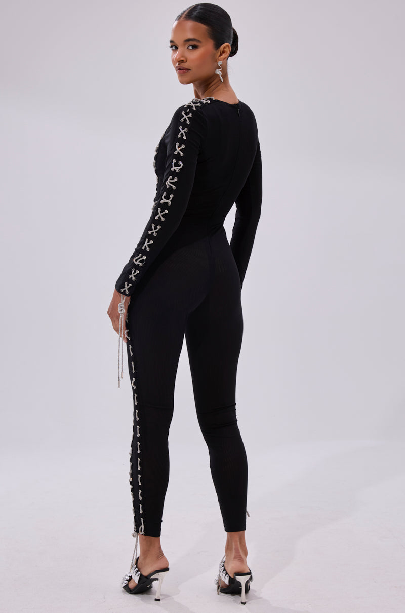 A woman in the LACE IT UP MESH JUMPSUIT WITH RHINESTONE DETAIL, featuring fitted black mesh with white X patterns, stands facing away and looks over her shoulder, styled with heels, statement earrings, and slicked-back hair.