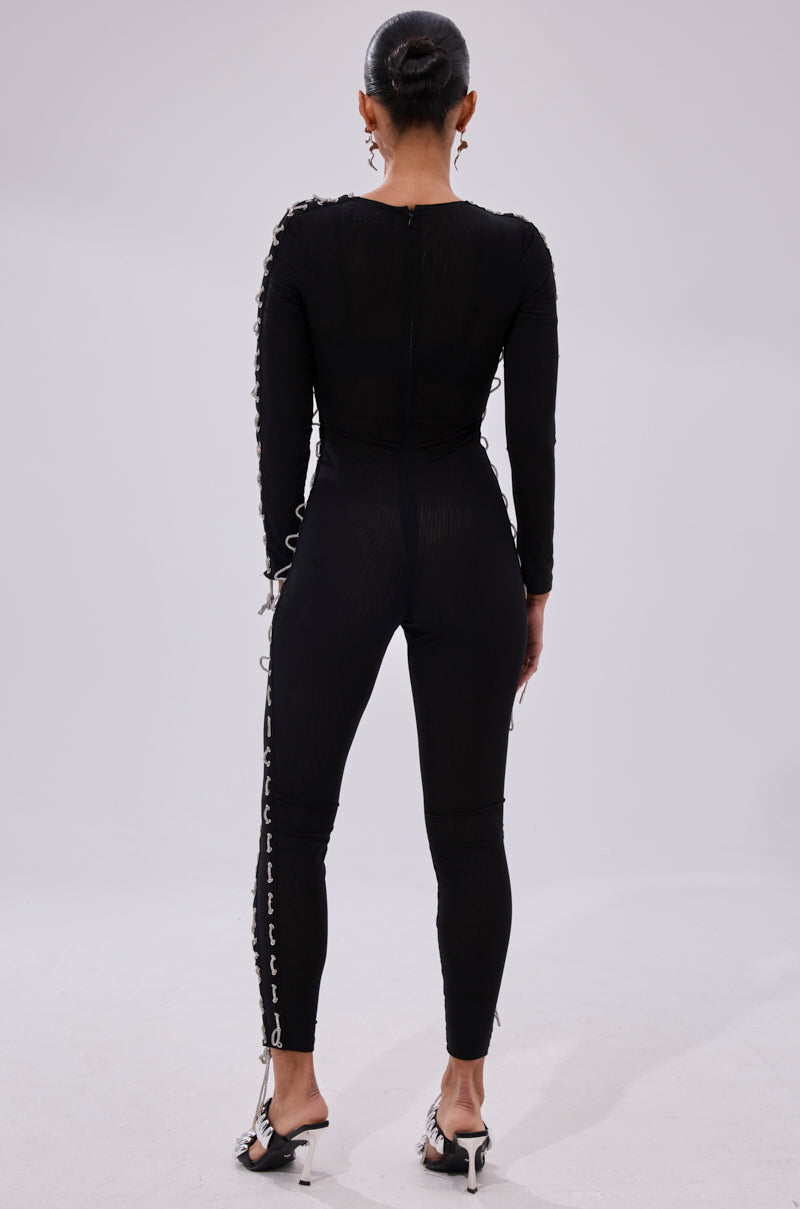 A person with hair in a neat bun stands facing away, wearing the LACE IT UP MESH JUMPSUIT WITH RHINESTONE DETAIL featuring crisscross lace-up accents on the arms and legs, paired with white heeled sandals.