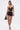 A woman in a black leather strapless corset top, the LOOK AT THIS ULTRA MINI SKIRT, strappy high-heeled sandals, and holding a small black handbag stands against a plain white background.
