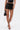 A woman stands against a plain white background wearing the LOOK AT THIS ULTRA MINI SKIRT in black faux leather with lace-up details, paired with a black top, strappy high-heeled sandals, and holding a small black handbag.