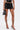 A person models the LOOK AT THIS ULTRA MINI SKIRT in black faux leather with lace-up details, paired with black strappy high heels and a small black handbag, standing against a plain background.