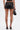 A person is seen from the back wearing the LOOK AT THIS ULTRA MINI SKIRT in black faux leather with lace-up details and long ties, paired with black strappy high-heeled sandals and a small black handbag.
