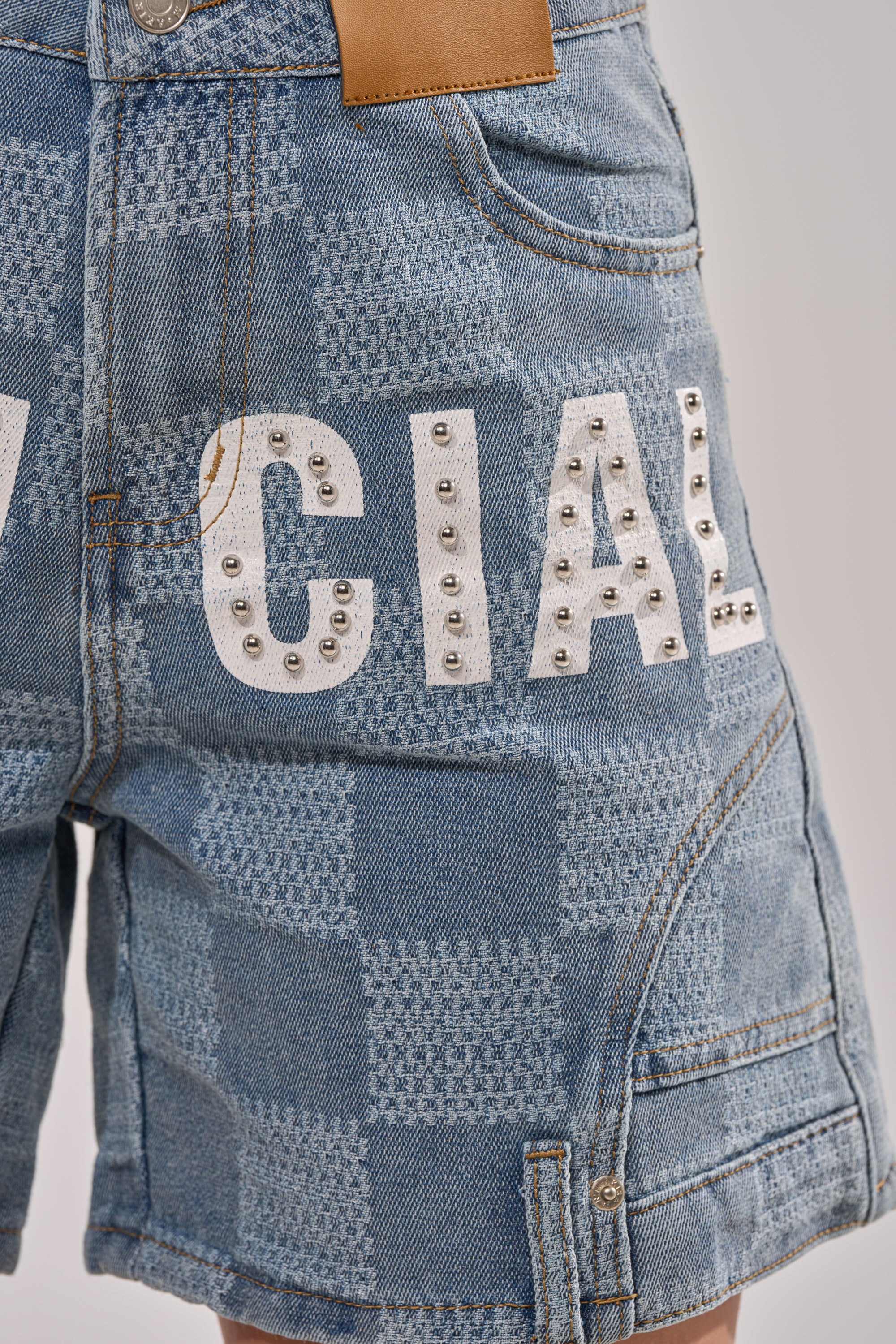 Close-up of the OFFICIAL DENIM BERMUDA SHORT in light-wash denim, featuring a checkered pattern, white printed letters, metal studs, multiple pockets, and a tan leather patch on the waistband. 8