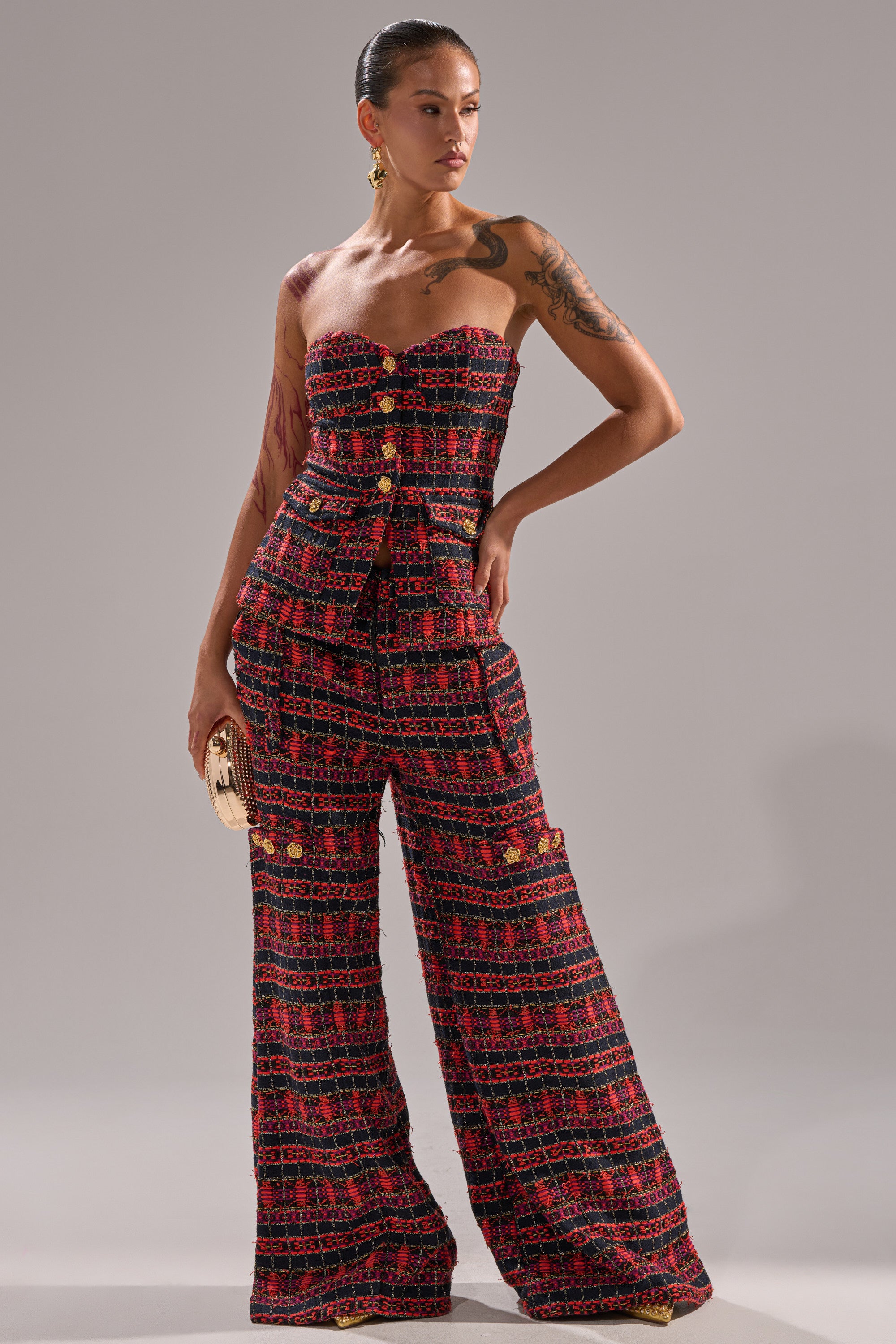 A person poses confidently against a plain backdrop, wearing the ONLY THINK ABOUT YOU TROUSER IN TWEED, styled with gold buttons, vintage wide-leg pants, and hoop earrings, while holding a small clutch purse.