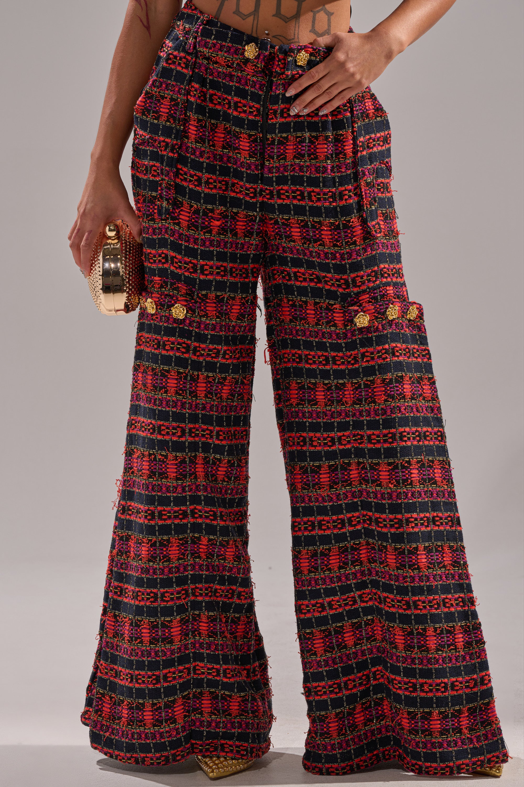 A person stands wearing ONLY THINK ABOUT YOU TROUSER IN TWEED, featuring high-waisted, wide-leg tweed pants with a red and black geometric pattern, vintage flair, and holding a small gold clutch. Torso and shoes are partially visible. 2
