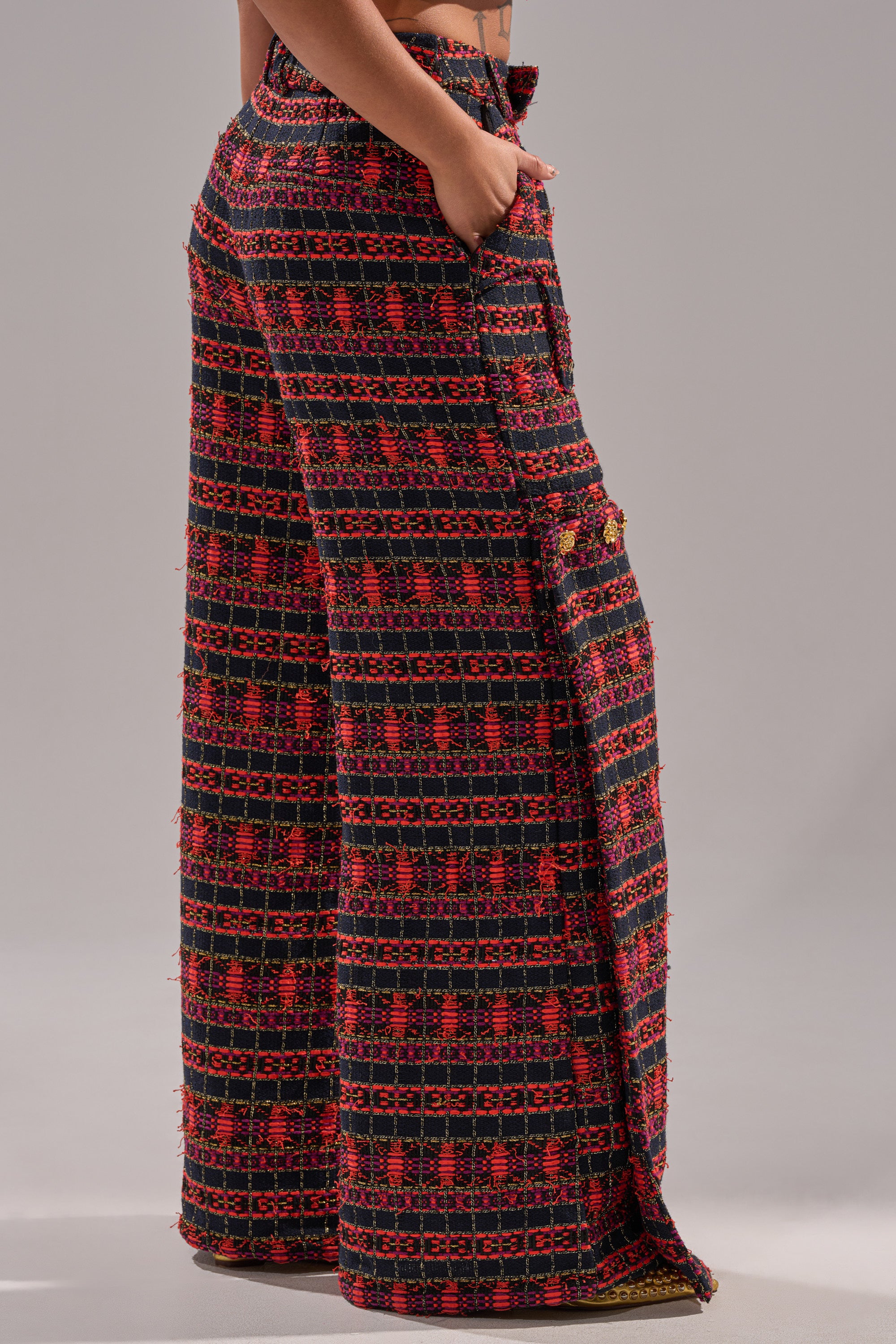 A person stands with hands in pockets, wearing ONLY THINK ABOUT YOU TROUSER IN TWEED—high-waisted, wide-leg pants featuring textured fabric with black, red, and orange geometric patterns—creating a perfect vintage girl fashion vibe against a neutral background. 3