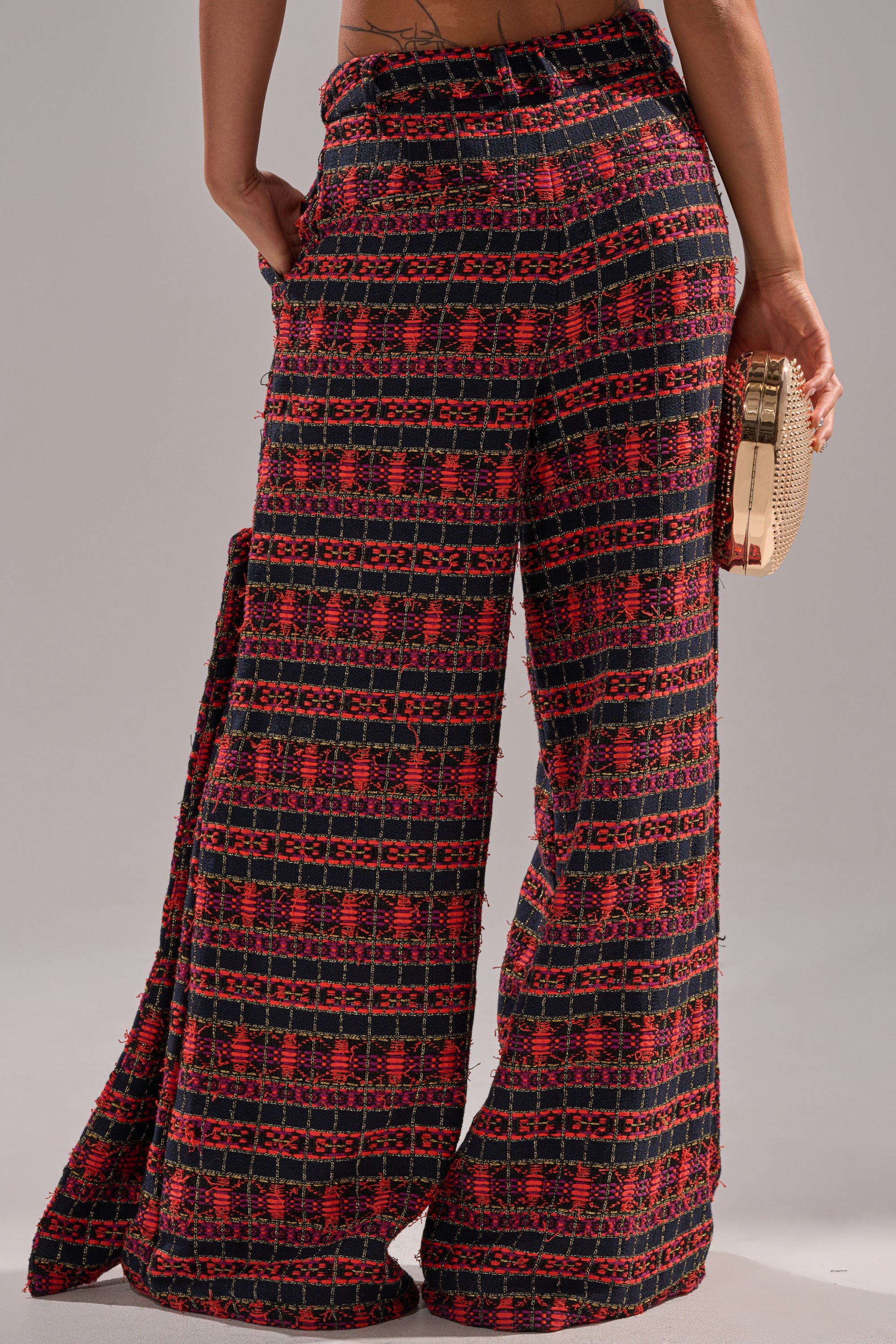 Wearing the ONLY THINK ABOUT YOU TROUSER IN TWEED—high-waisted, wide-leg pants with a red and black geometric pattern—a person stands sideways, vintage style, holding a textured beige clutch with one hand in their pocket. 4