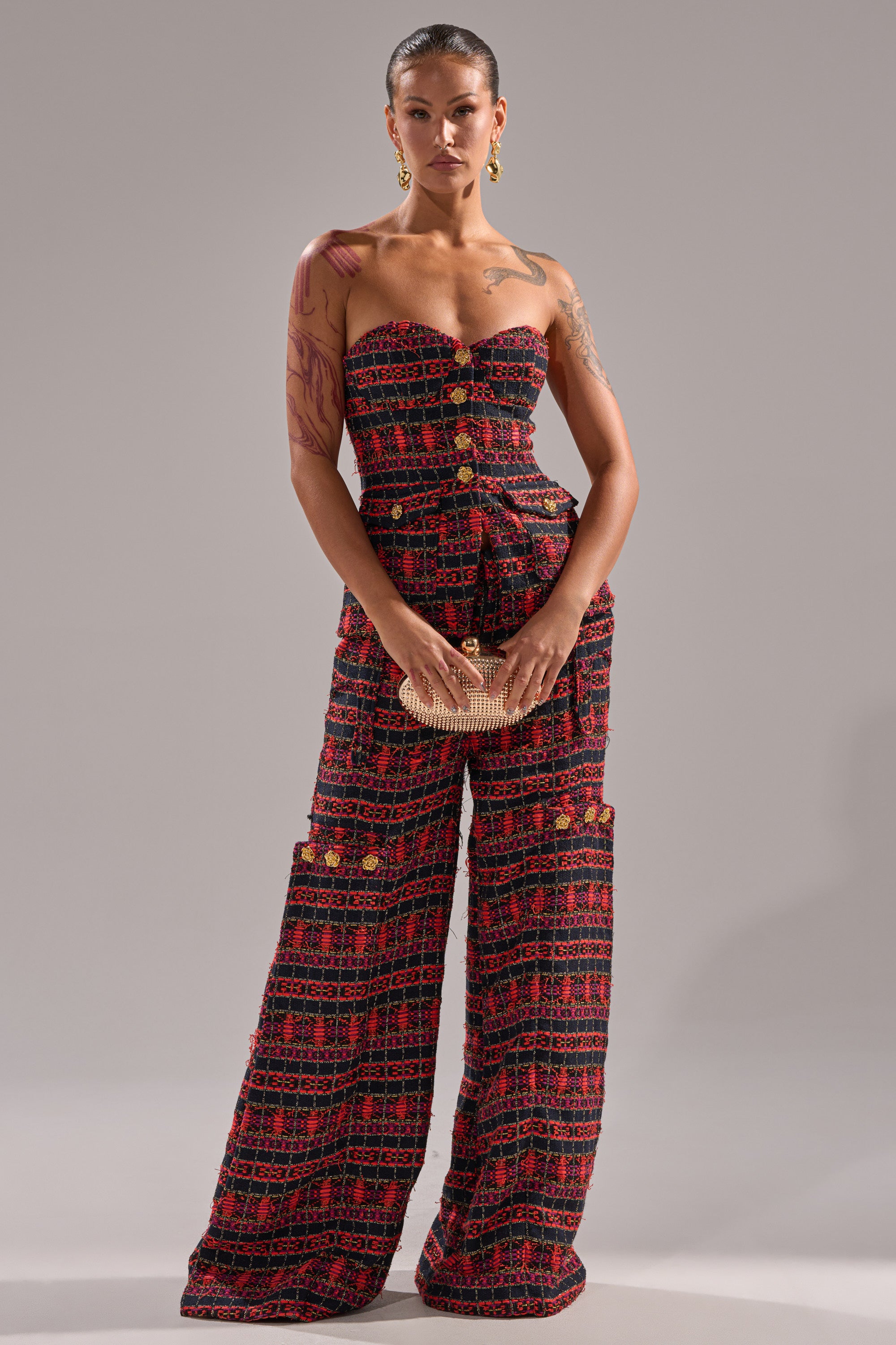A vintage girl stands against a plain backdrop, wearing the ONLY THINK ABOUT YOU TROUSER IN TWEED, a wide-leg red and black patterned jumpsuit. She accessorizes with gold earrings, a beige clutch, and displays tattoos on her arms. 5