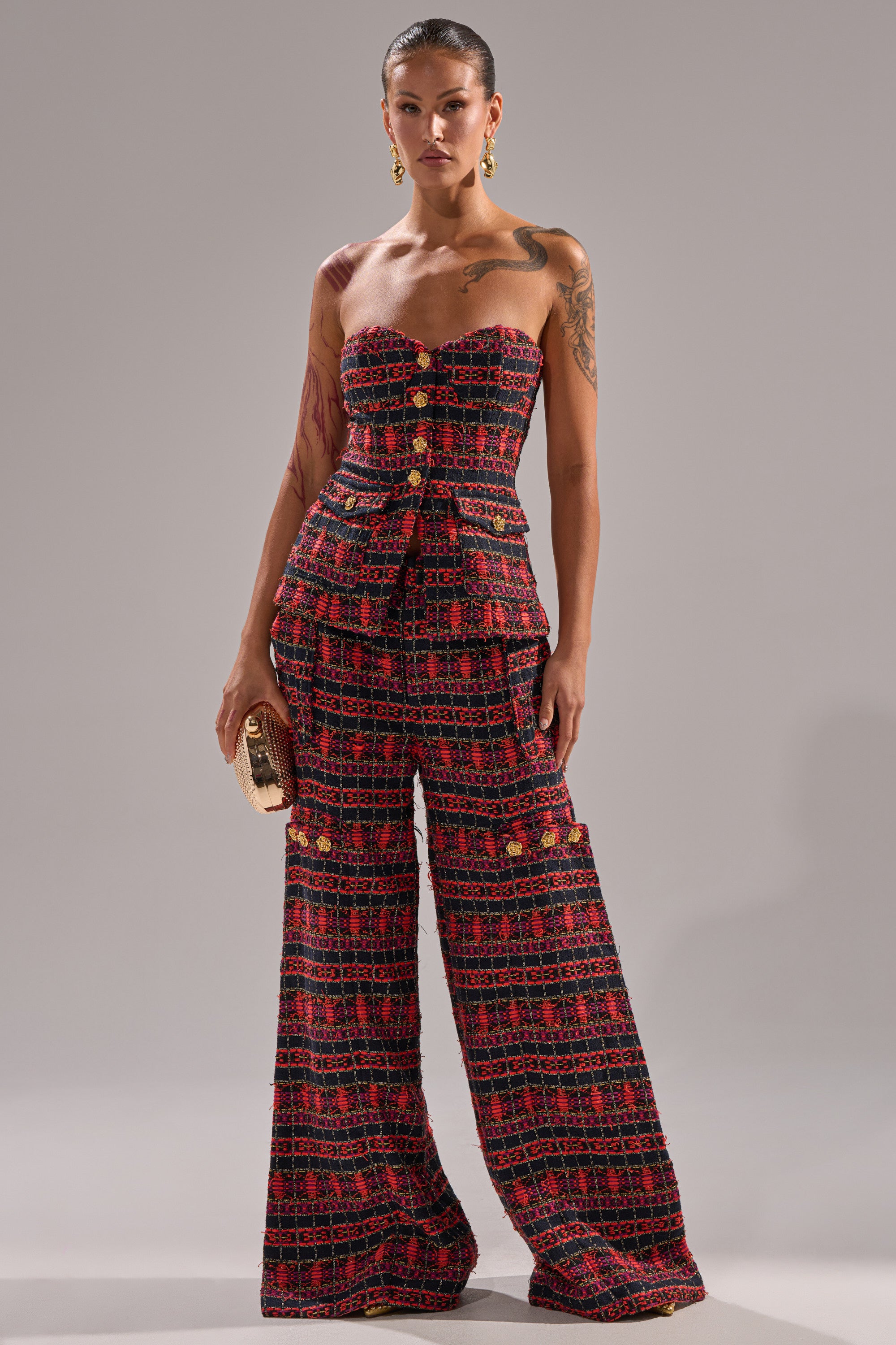 A woman wears the ONLY THINK ABOUT YOU TROUSER IN TWEED—a strapless, fitted red and black plaid jumpsuit with gold buttons and wide-leg pants. She accessorizes with a clutch, gold earrings, arm tattoos on display, and her hair pulled back. 6