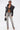 A person wearing a black leather jacket, sheer black bralette, ULTRA DISTRESSED SKINNY DENIM jeans, high-heeled sandals, sunglasses, and holding a black handbag stands confidently against a plain white background.