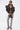 A stylish person poses confidently against a white background wearing a structured black leather jacket, black bralette, ULTRA DISTRESSED SKINNY DENIM, black high heels, sunglasses, and carrying a matching handbag.