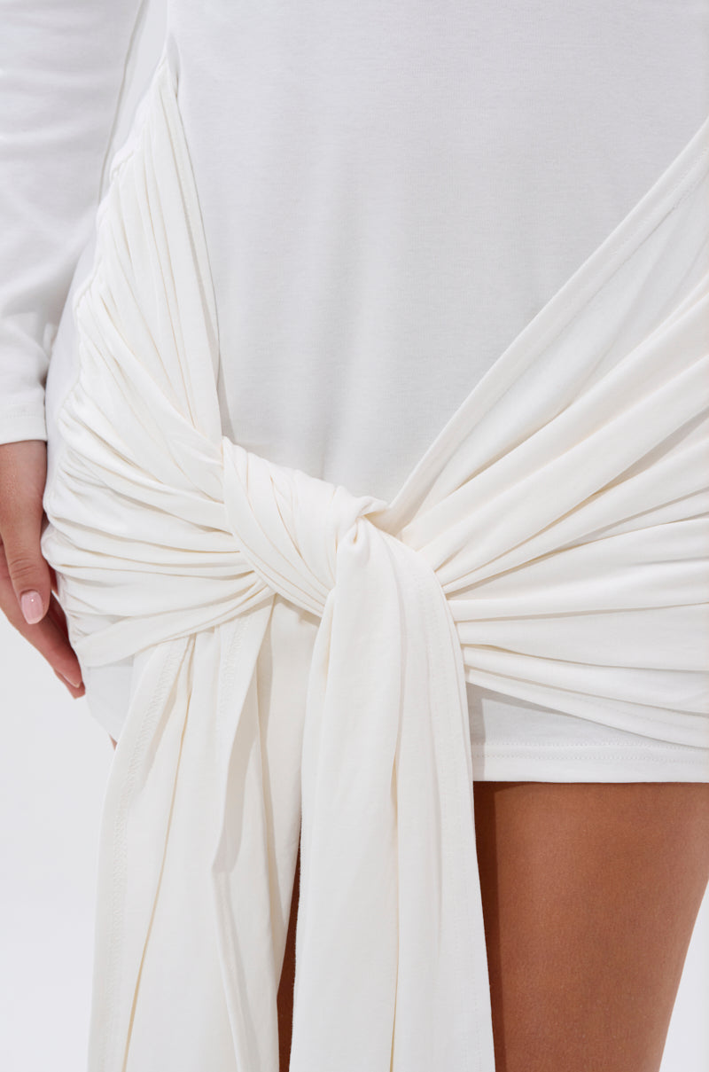 A close-up of the SIMPLE GIRL TIE-FRONT T-SHIRT DRESS in white, featuring long sleeves and a large draped knot detail at the hip with flowing fabric, worn by a light-skinned model.