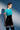 A woman with short brown hair stands against a blue and white background, wearing a fitted turquoise top, thigh-high patterned stockings, and the LETS DO BUSINESS MINI SKIRT. She looks confidently to the side with a soft shadow behind her.