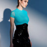 A woman with short brown hair stands against a blue and white background, wearing a fitted turquoise top, thigh-high patterned stockings, and the LETS DO BUSINESS MINI SKIRT. She looks confidently to the side with a soft shadow behind her.
