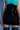 A person models the LETS DO BUSINESS MINI SKIRT in shiny patent faux leather, paired with a turquoise sleeveless top and black patterned thigh-high stockings, shown from shoulders to mid-thigh against a blue background.