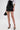 A person wears a white blouse, the LETS DO BUSINESS MINI SKIRT in patent faux leather with a high-rise waist, black heels with gold accents, a gold bracelet, and holds a small black handbag with a gold handle. The background is plain white.