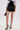 Wearing the LETS DO BUSINESS MINI SKIRT, a shiny patent faux leather mini with a high-rise waist, paired with black pointed-toe heels, a white blouse, gold jewelry, and holding a black handbag against a plain background.