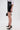A person wears a white shirt, the LETS DO BUSINESS MINI SKIRT—a high-rise patent faux leather mini skirt—black heels with gold accents, a chunky gold bracelet, and holds a small black handbag against a plain white background.