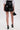 A person faces away, wearing the LETS DO BUSINESS MINI SKIRT in shiny black faux leather with a high-rise waist and lace-up detail, paired with a white blouse, black heels, gold jewelry, and holding a small black handbag.