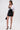 A woman stands against a plain background wearing a white off-shoulder shirt, visible black lace bra, LETS DO BUSINESS MINI SKIRT in patent faux leather, black heels with gold accents, gold earrings, chunky gold bracelet, and holding a small black bag.