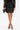 A woman wearing the THIS IS THE LIFE FAUX LEATHER MINI SKIRT, a black lace top, black pointed-toe heels with gold accents, and holding a black feathered clutch stands against a white background.