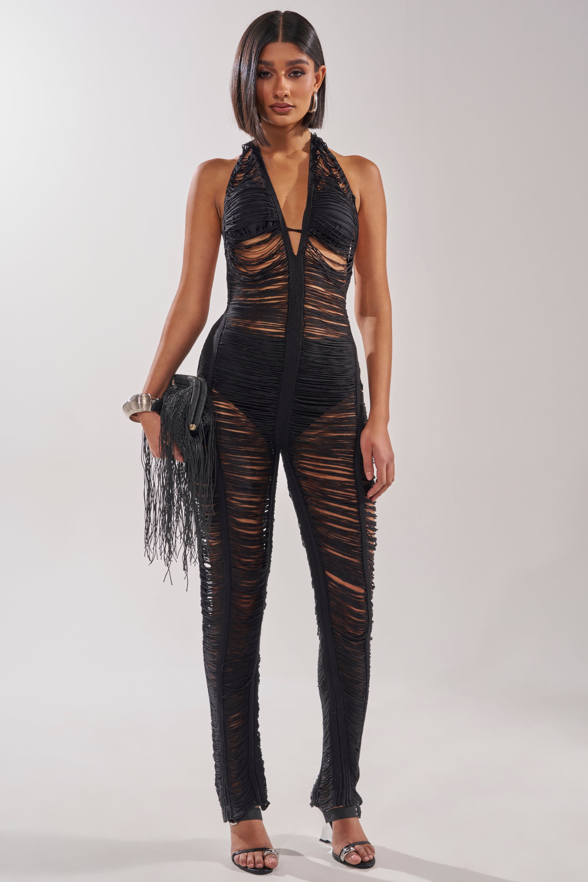 A woman faces forward in the IT GIRL SHREDDED JUMPSUIT, a sleeveless, sheer black jumpsuit with ripped details. She completes her edgy festival look with a fringed clutch, high-heeled sandals, and a sleek bob. 1