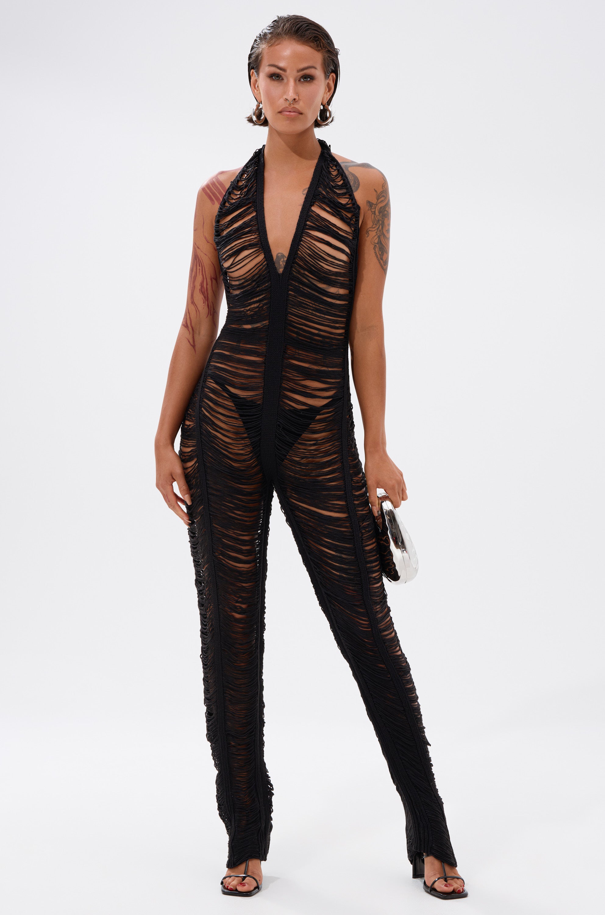 A woman exudes confidence in the IT GIRL SHREDDED JUMPSUIT—a sheer, black, textured halter piece styled with visible black underwear. She accessorizes with a silver bag, black heeled sandals, hoop earrings, and showcases tattoos and wet-look hair.