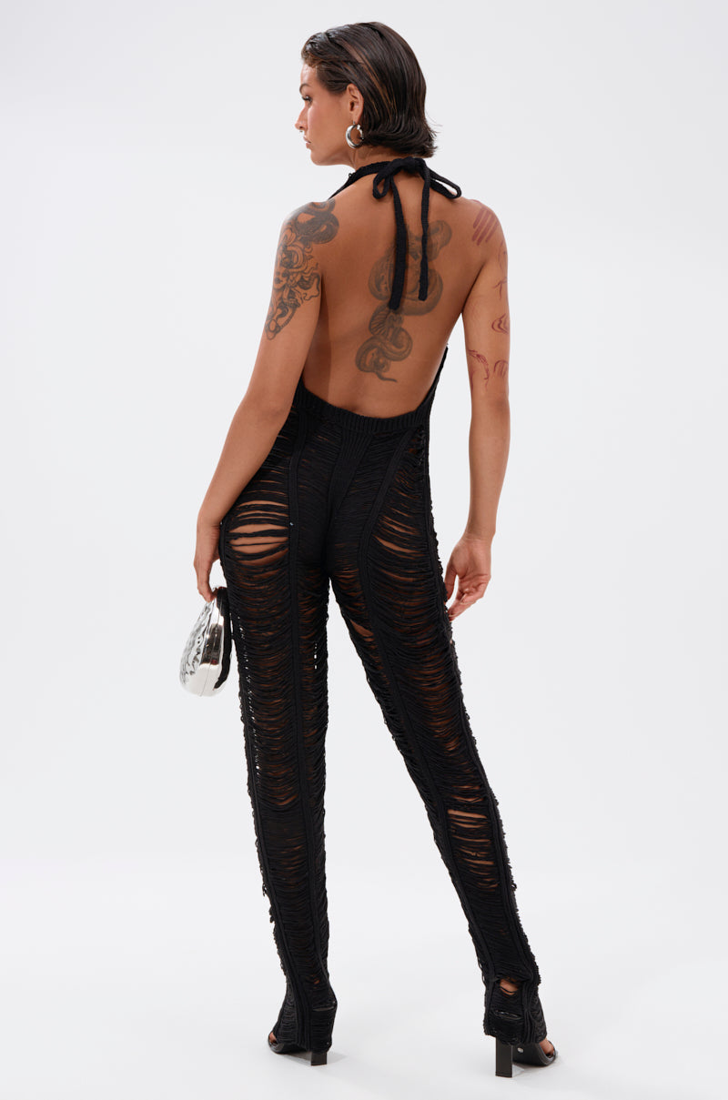 A person with wet, slicked-back hair and tattoos models the IT GIRL SHREDDED JUMPSUIT—a curve-hugging, backless halter style with black sheer shredded details—paired with heels and a metallic handbag against a white background.