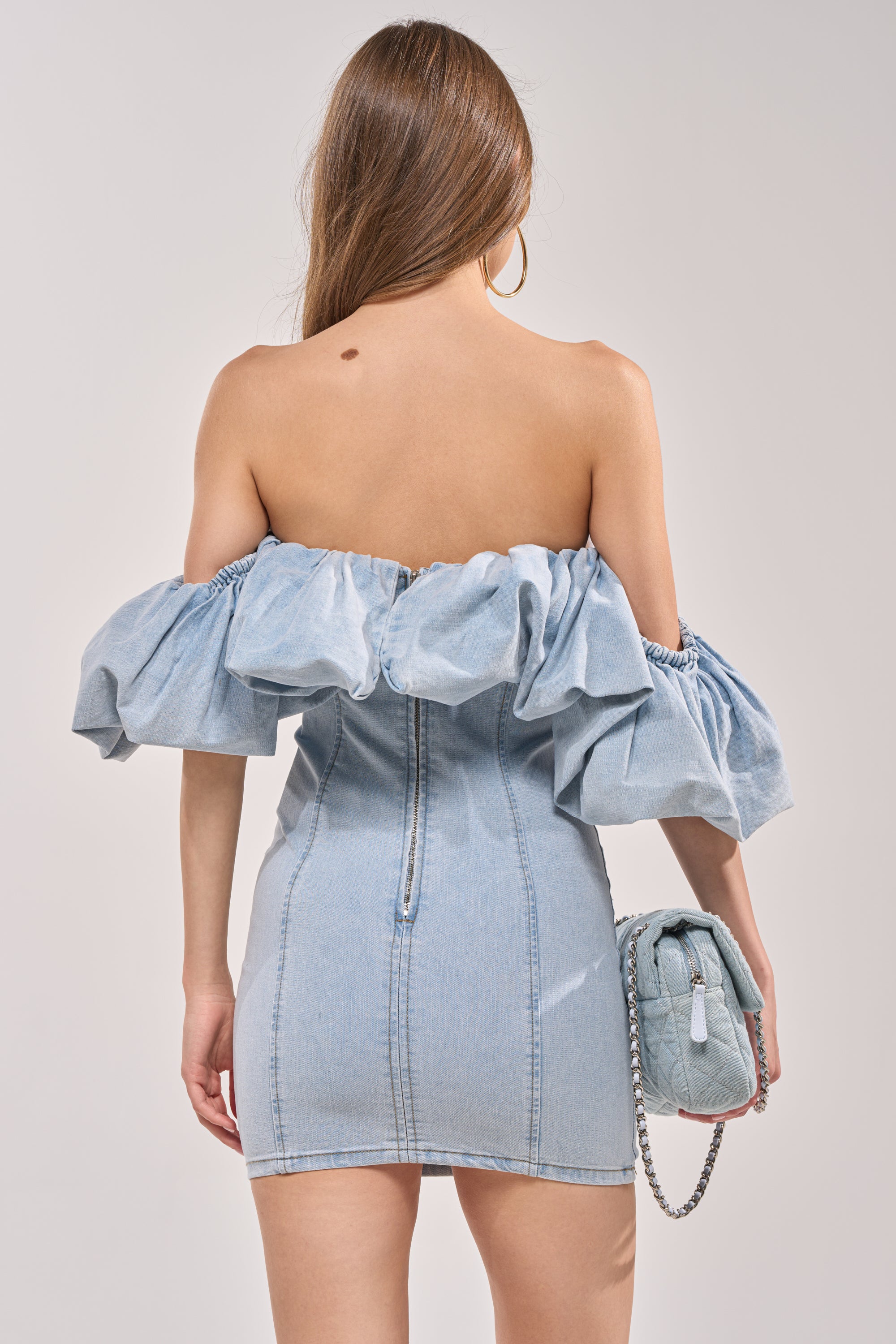 A woman with long brown hair is seen from the back, wearing the JULIA DENIM MINI DRESS off the shoulder, styled with a matching denim purse and large hoop earrings. 3