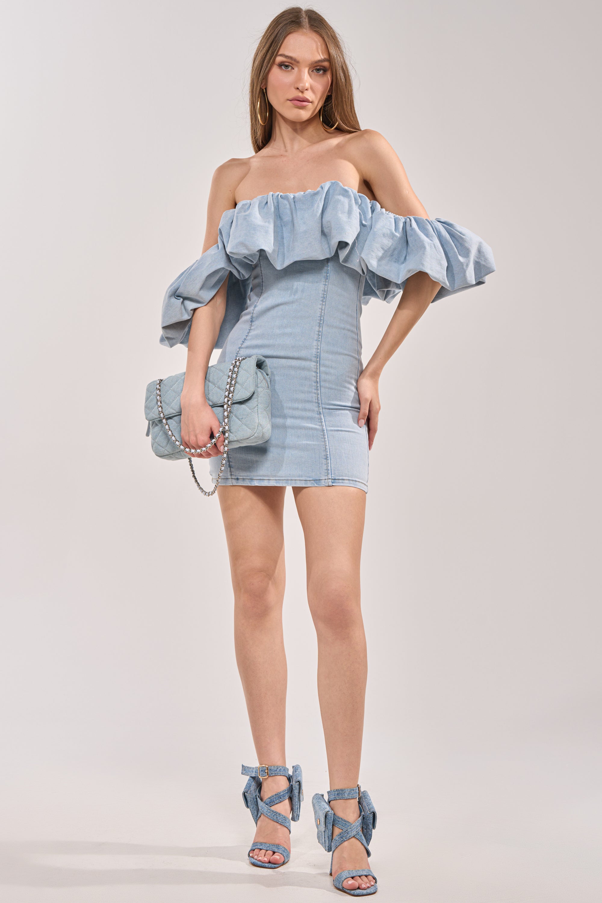 A woman wears the JULIA DENIM MINI DRESS—strapless, ruffled, and light blue—paired with matching denim sandals and a quilted denim handbag, standing against a plain light background. 4