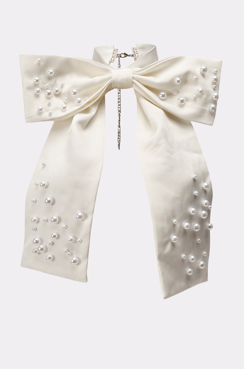 The TAKE ME ANYWHERE BOW NECKLACE features a large, elegant white bow with pearl accents and a silver chain clasp—an eye-catching oversized choker ideal as a bridal necklace or statement piece. Shown on a simple light background. 2