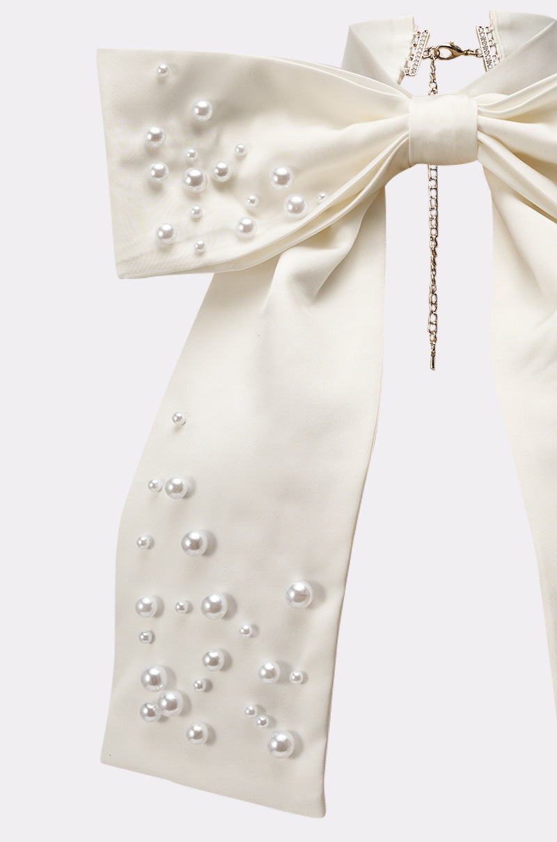 The TAKE ME ANYWHERE BOW NECKLACE features an oversized ivory bow with a chain clasp, adorned with small, medium, and large pearls scattered along one trailing ribbon end. 3