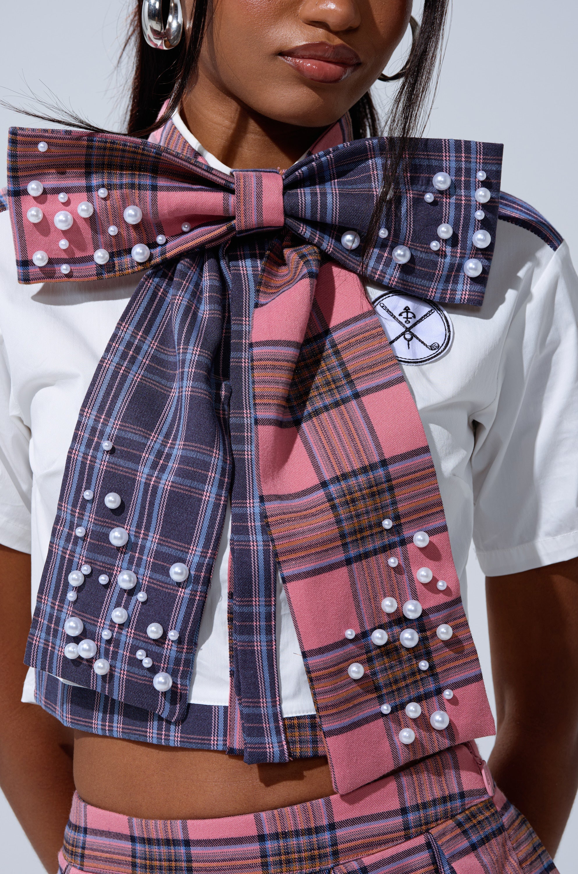 A woman styles a white crop top with an embroidered patch and a plaid bow tie accented with faux pearls, paired with a matching high-waisted plaid skirt. The look is finished with silver hoops and the DREKE OVER THE TOP BOW NECKLACE.