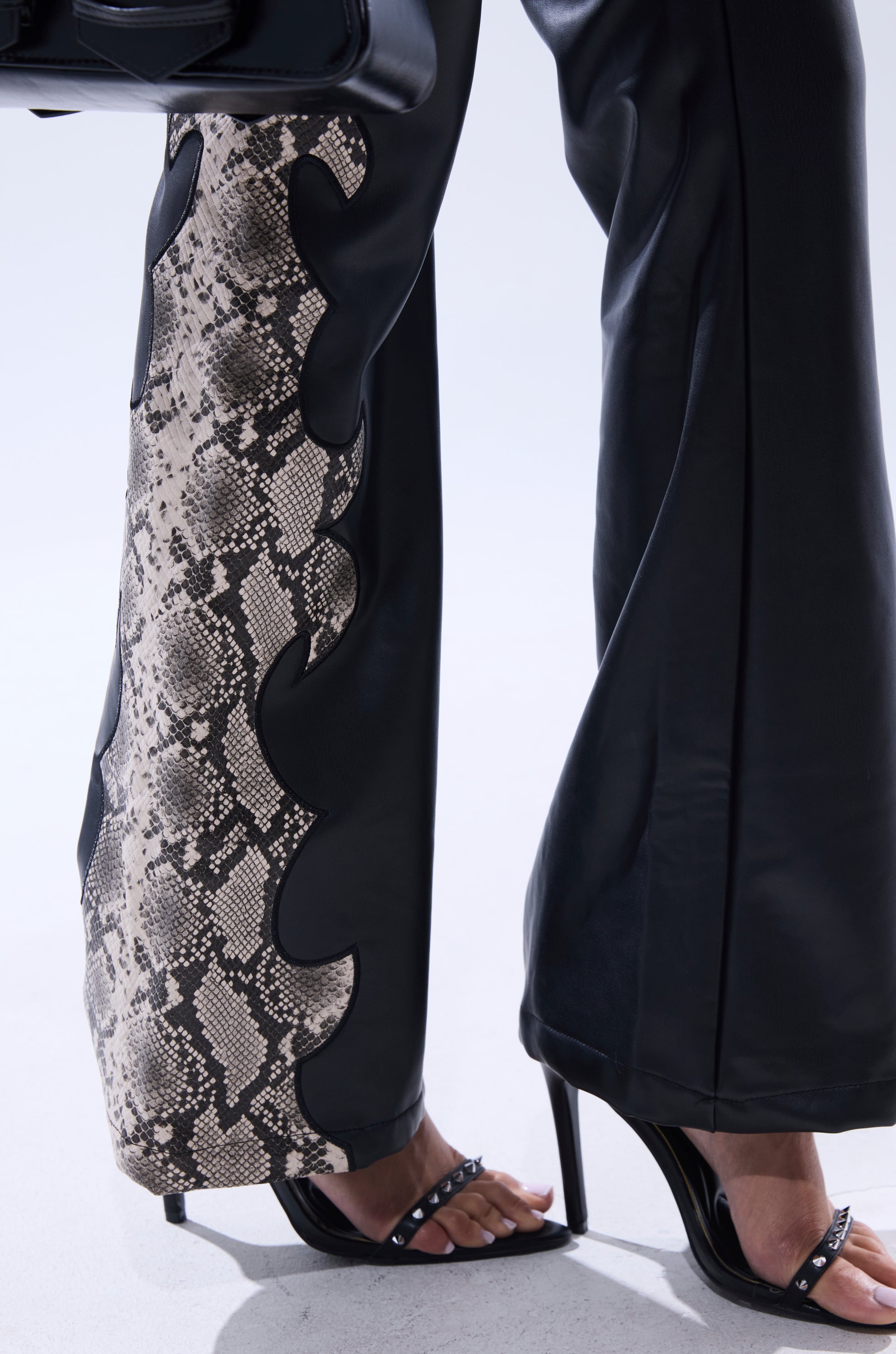 A person stands on a light surface, showing only their legs and feet in black high-heeled sandals and VENOMOUS FAUX LEATHER PANTS featuring a bold snakeskin print panel on the side.