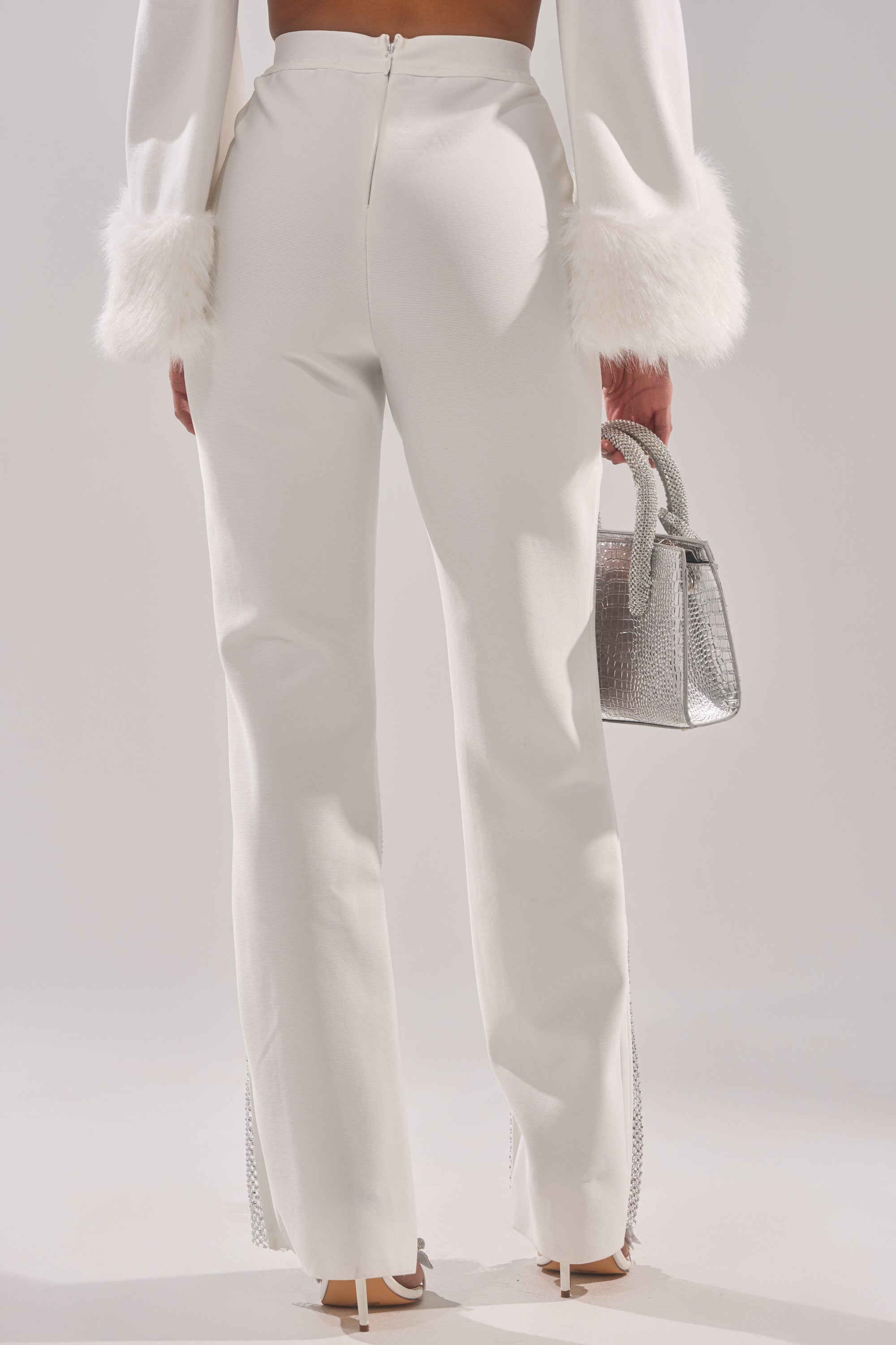 Someone stands facing away, wearing the PALMER BANDAGE TROUSER WITH RHINESTONE MESH—white high-rise pants with split hems and a zip closure—paired with a matching white top, beige heels, and a silver rhinestone mesh handbag.