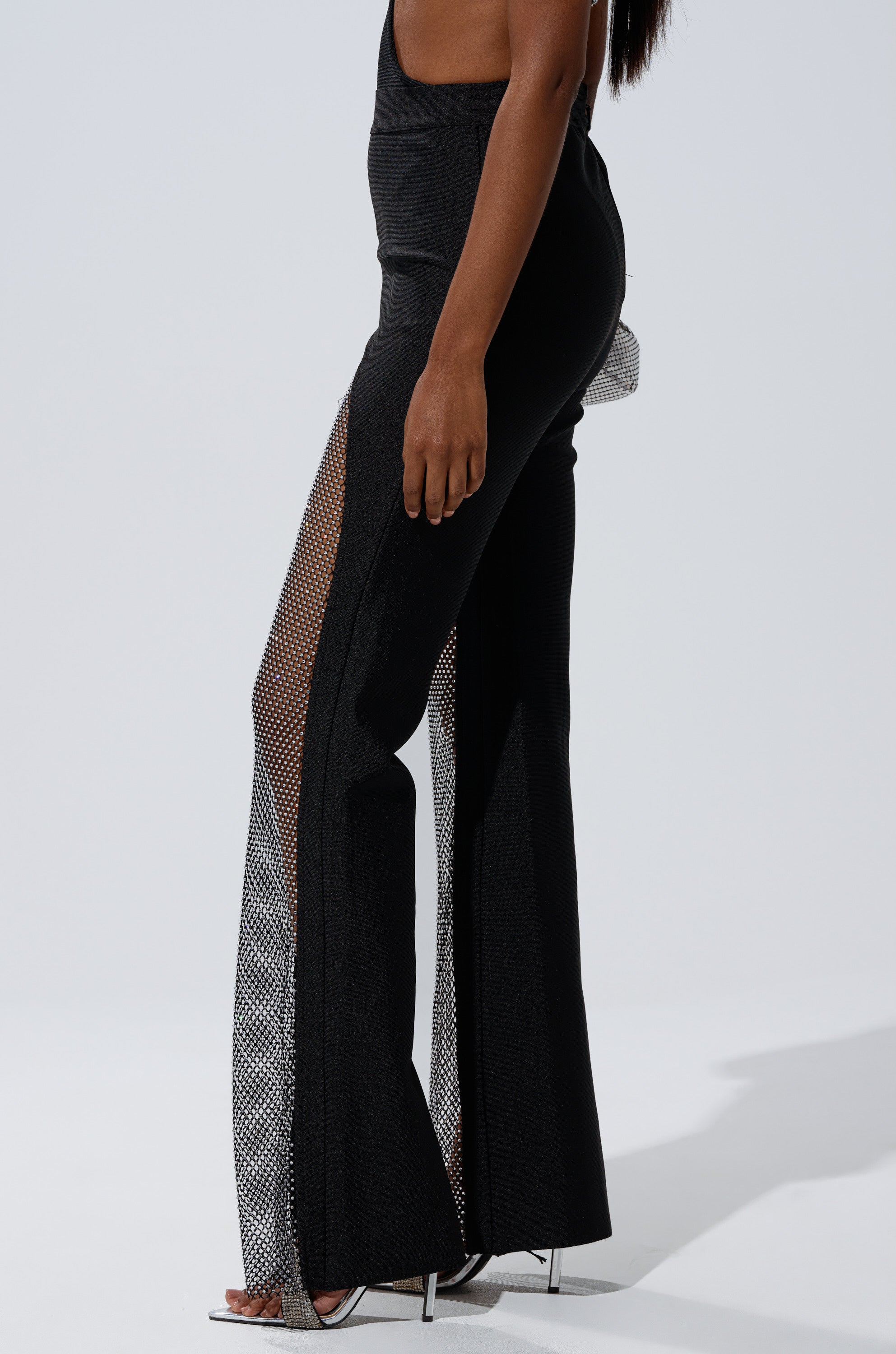 A person models the JULIA BANDAGE TROUSER WITH MESH RHINESTONES—black, high-waisted pants featuring mesh rhinestone side panels—paired with silver heels against a plain light gray background, cropped above the torso.