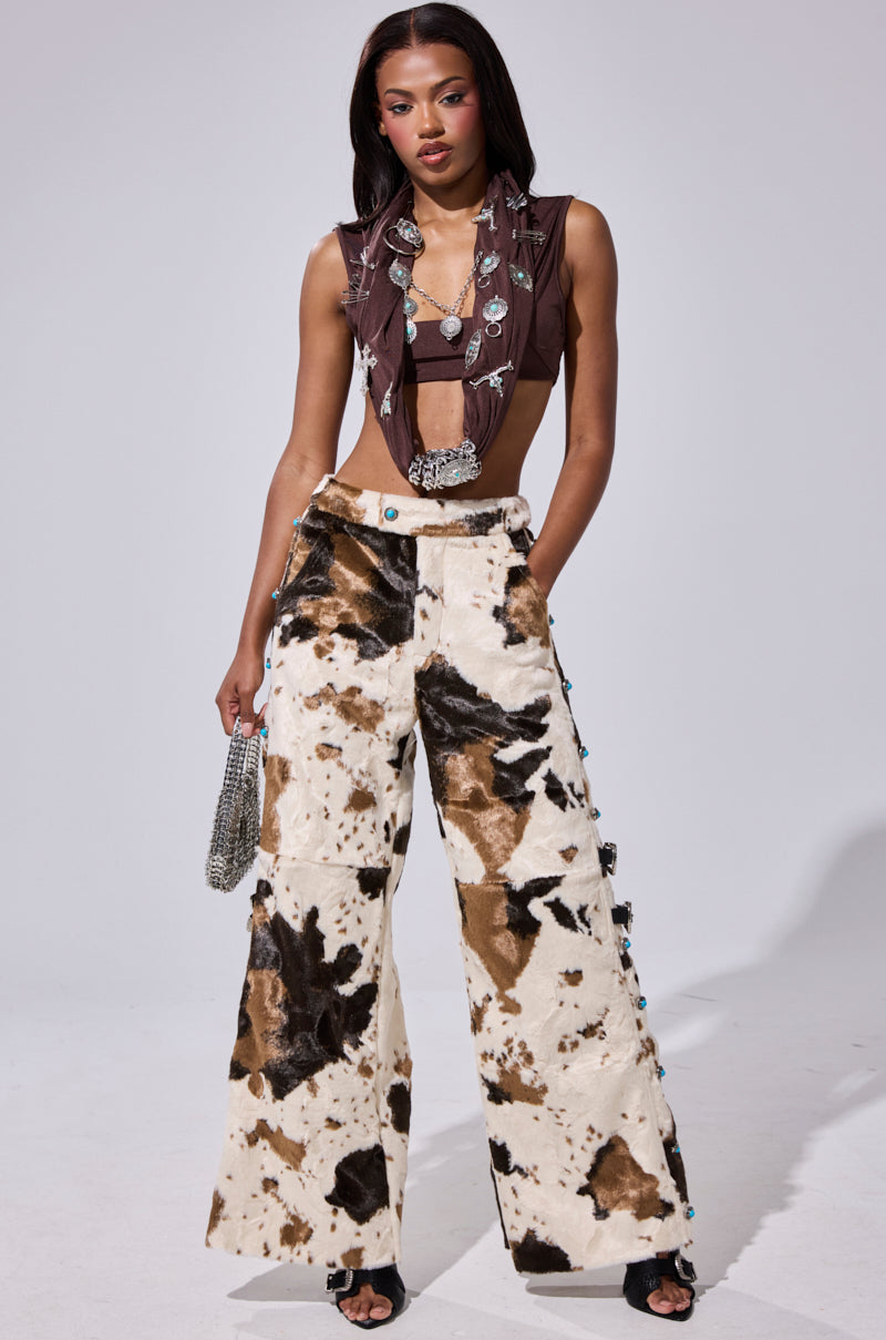 A woman stands confidently in HIGHLAND COW FAUX FUR PANT, a brown and white cow-print wide-leg style, paired with a brown sleeveless crop top with silver details, black sandals, and a small silver handbag.