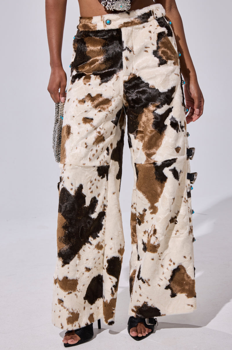 A person shows off cowgirl style in HIGHLAND COW FAUX FUR PANT—wide-leg, high-waisted pants with a brown and black cow print, side button details—styled with black open-toe heels and a chain accessory against a plain white background.
