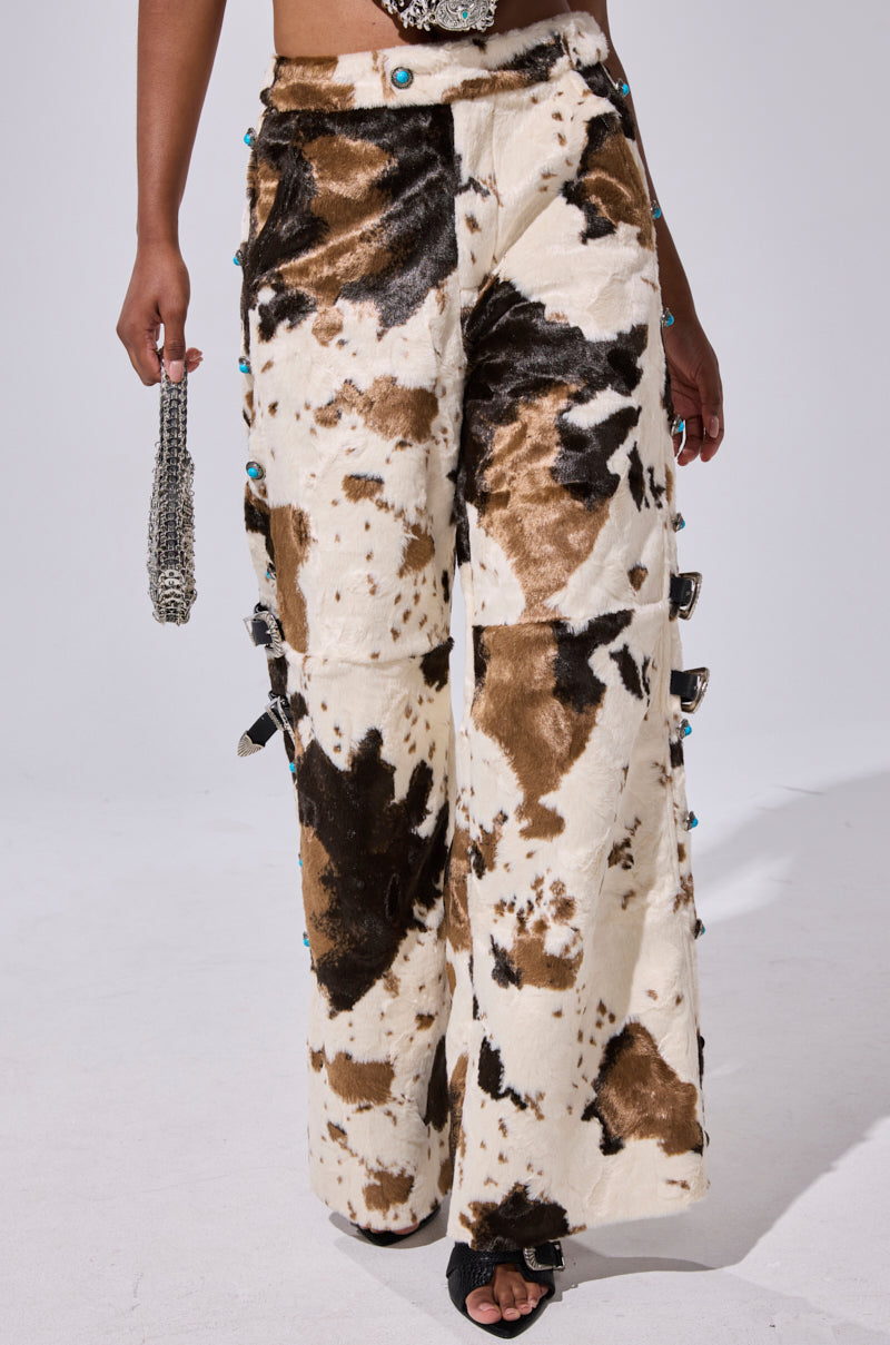 Wearing the HIGHLAND COW FAUX FUR PANT with a bold cow print, blue accents, and side buckles, this look channels cowgirl flair, paired with a silver handbag and black open-toe heels.