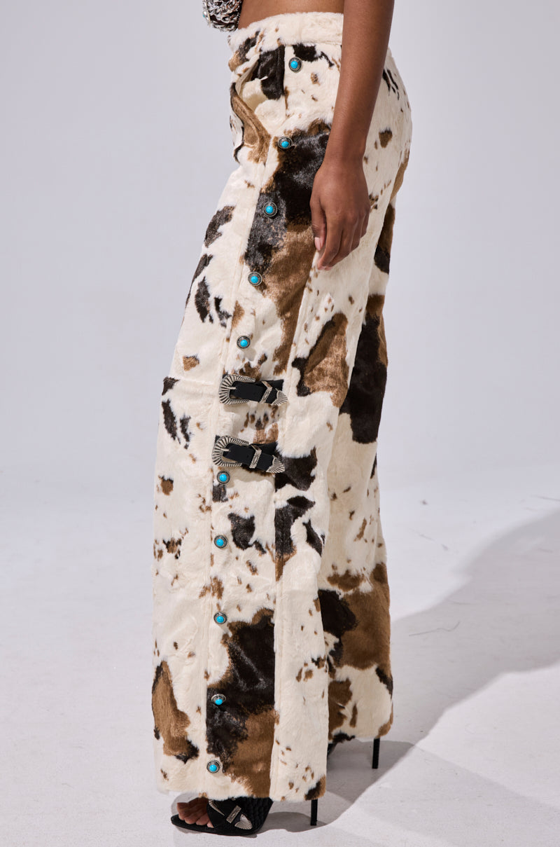 A person wears the HIGHLAND COW FAUX FUR PANT—wide-leg pants with brown and black cow-print patches, turquoise buttons, and silver side buckles—paired with black high-heeled shoes.