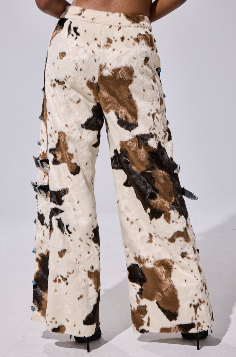 A person stands facing away from the camera against a plain background, wearing HIGHLAND COW FAUX FUR PANT—high-waisted, wide-leg pants featuring a cream and brown cow print pattern.