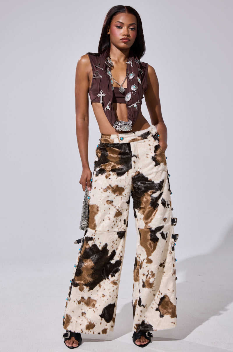A woman stands confidently in HIGHLAND COW FAUX FUR PANT featuring a brown and white cow print, paired with a sleeveless brown crop top with metallic accents, a small purse, and heeled sandals.