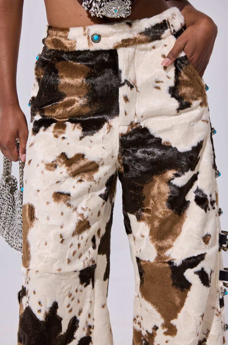 Someone models the HIGHLAND COW FAUX FUR PANT, featuring a wide-leg design in brown and white with turquoise buttons, paired with a silver chain-link handbag.