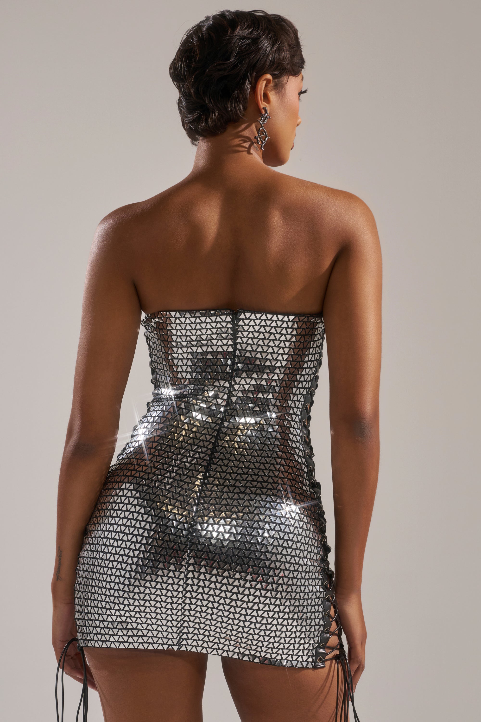 Seen from the back, a woman with short dark hair wears the MEET ME AT THE DISCO MINI DRESS—a silver, strapless mini dress with triangular patterns and side ties—for a chic, modern party look against a neutral background.